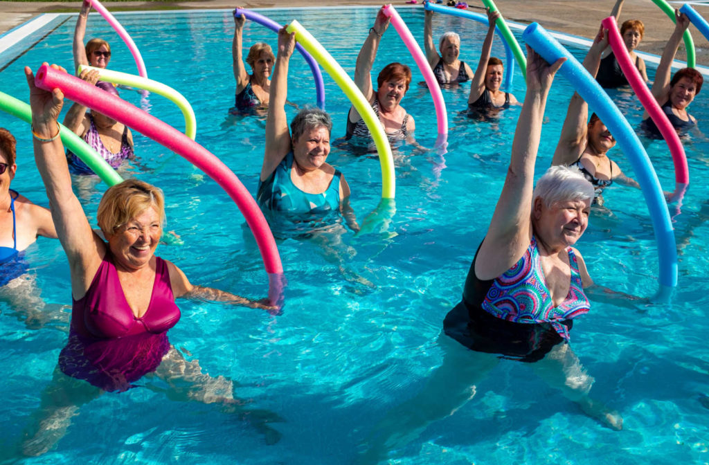 Elderly women exercising together with foam noodles in outdoor swimming pool.