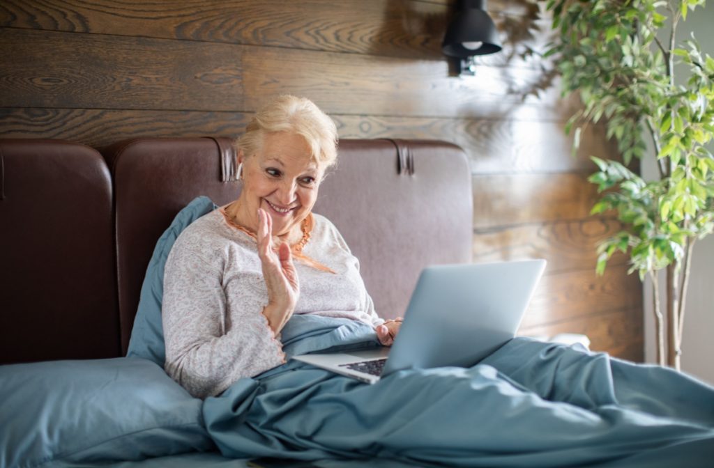A happy older adult woman, who is sitting on bed, in a video call with a loved one.