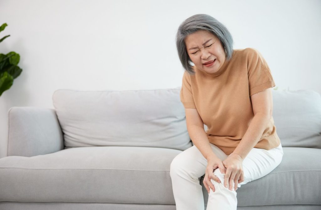 A senior woman sitting on a couch holds her knee in pain and winces