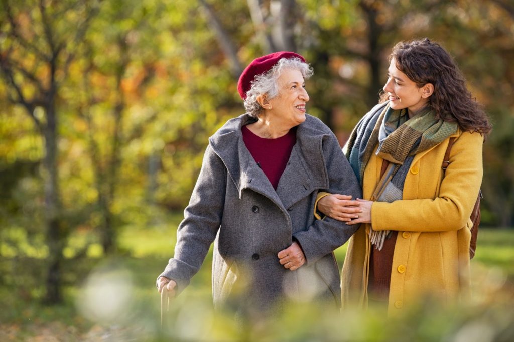 A young woman in a yellow coat threads her arm through an older woman’s while the both of them walk through a park and talk.