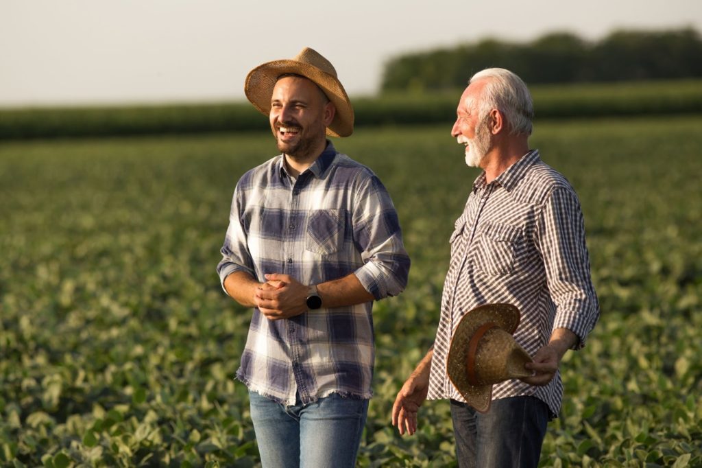 A young man and an older man speak in a farm field together. Both are wearing plaid shirts and have straw hats.