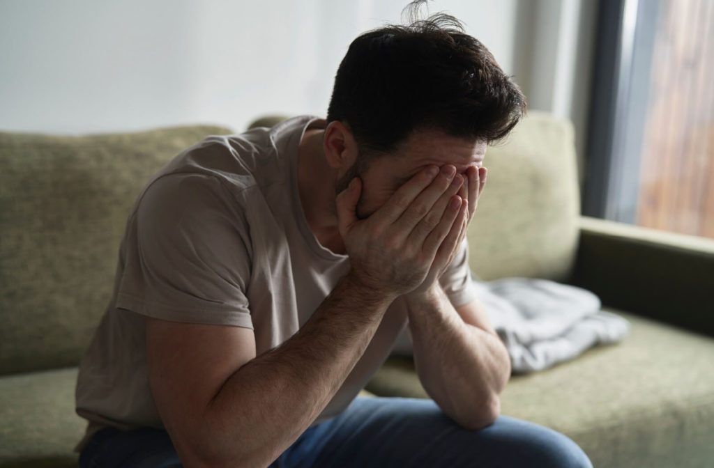 An exhausted male caregiver sitting alone on the couch with his head in his hands, trying to decide if it's time for respite care for his elderly parent.