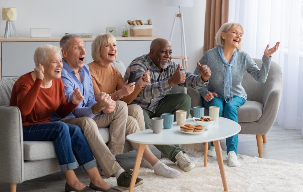 A group of seniors laugh and smile as they watch a show on a TV during a senior living social event.