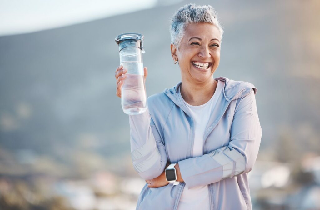 An older adult smiling while holding up a water bottle while out on a nature hike.