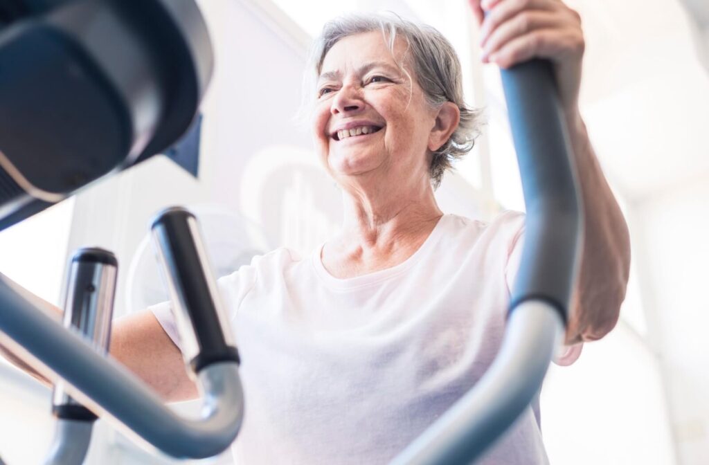 A senior smiles while using an elliptical in a fitness center offered by their senior living community, happy to be moving