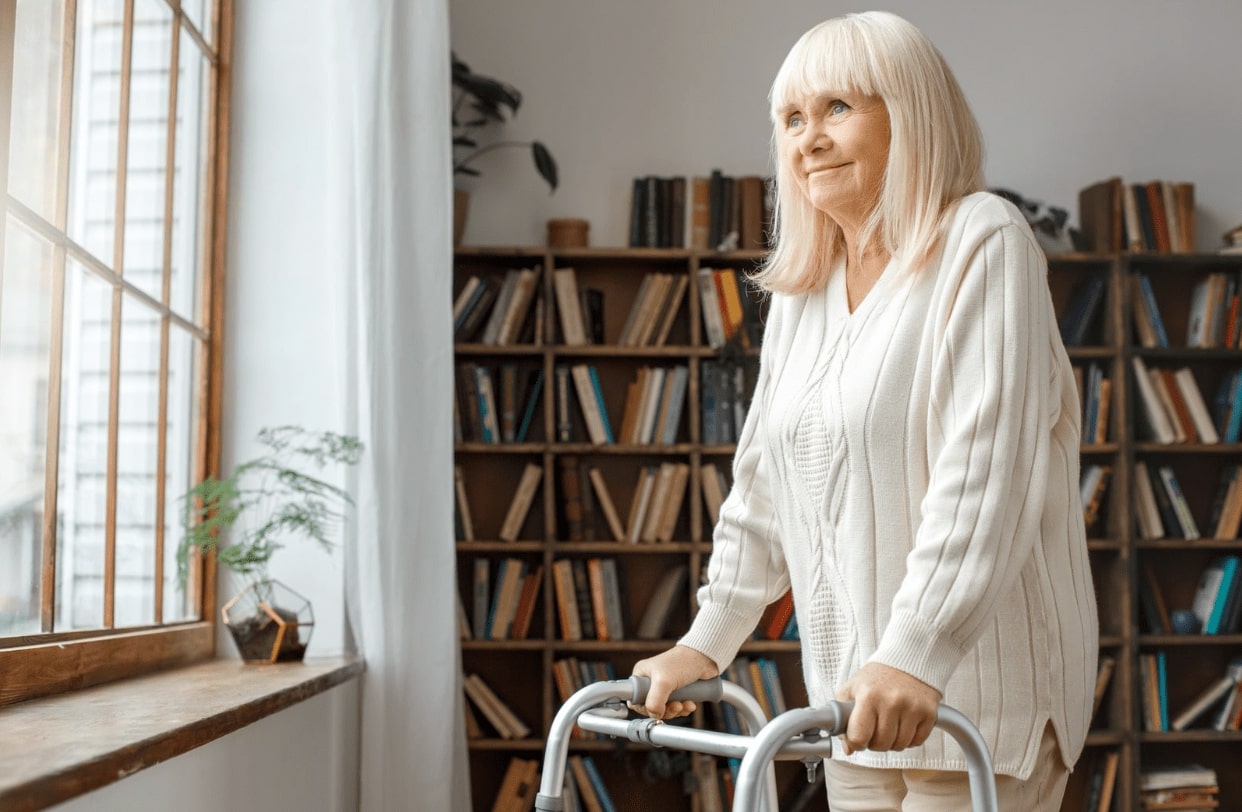 An older woman with a walker, looking out a window in an assisted living home.
