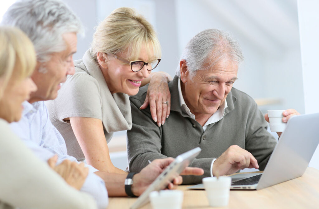 An older man and woman smiling while sitting at a table, drinking coffee and looking at a laptop. Another couple sits next to them, holding a tablet and looking on.